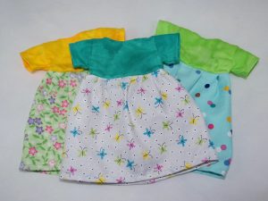 Three simple doll dresses. One is yellow with a green floral skirt, one is teal with a gray skirt covered in colorful butterflies, and one is green with a light blue skirt covered with colorful polka dots.